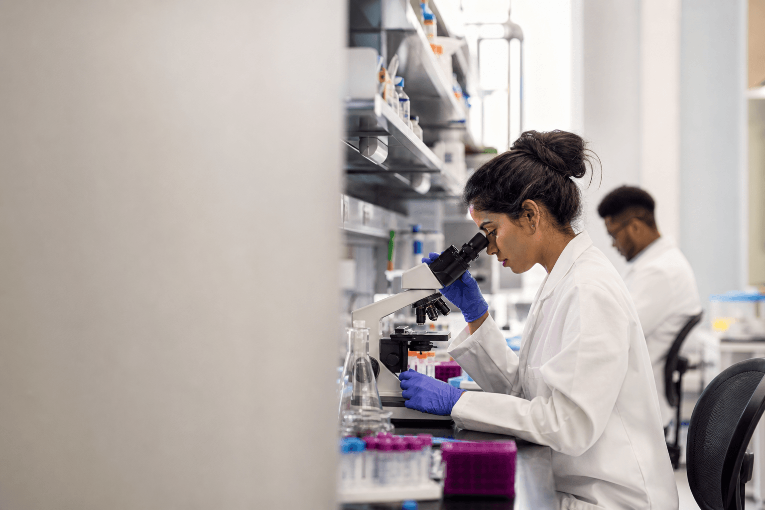 A graduate researcher in a white coat using a microscope at a molecular-biology bench lined with sample vials.