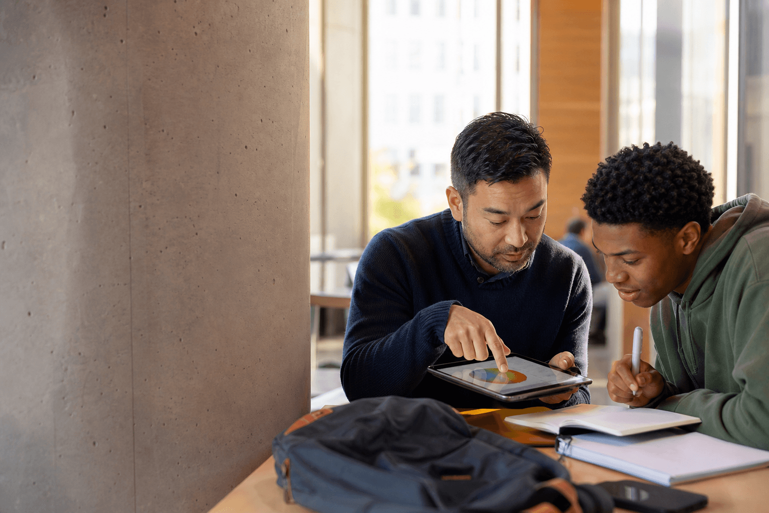 A tutor and a college student leaning over a tablet together in a bright study nook, discussing a diagram.